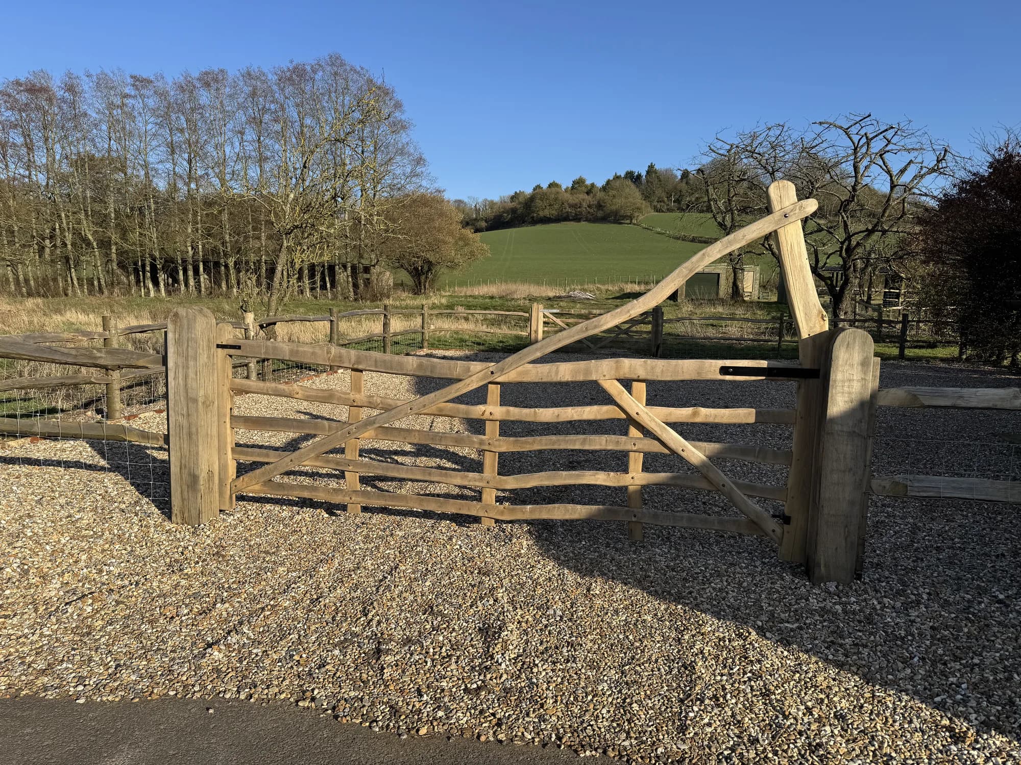 A curved rising-top five-bar gate we made, hung across a gravel drive with the Kent hills behind